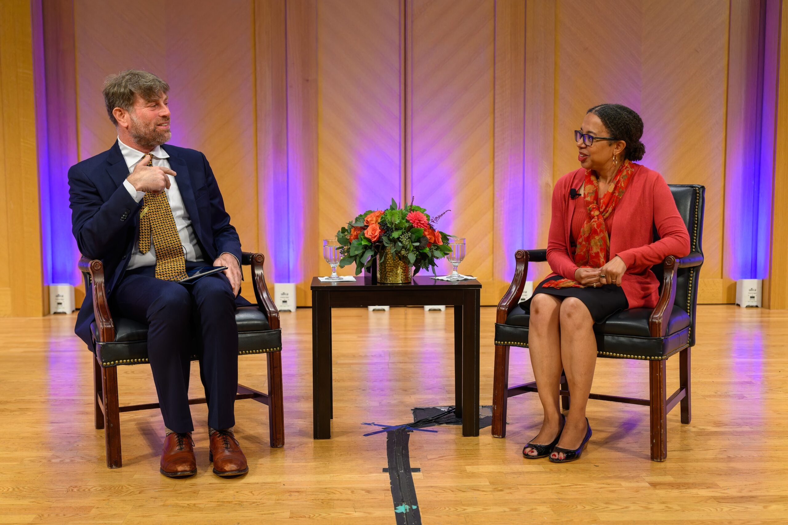 Adam Rothman, a white man wearing a navy suit, and Rachel Swarns, a black woman wearing a red and black skirt suit, are seated on a stage, angled toward each other and engaged in conversation.