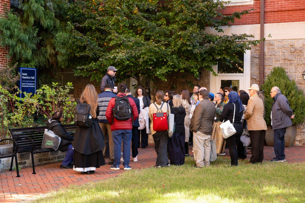 A white man in a baseball cap and sunglasses speaks to a large tour group outside of Isaac Hawkins Hall.