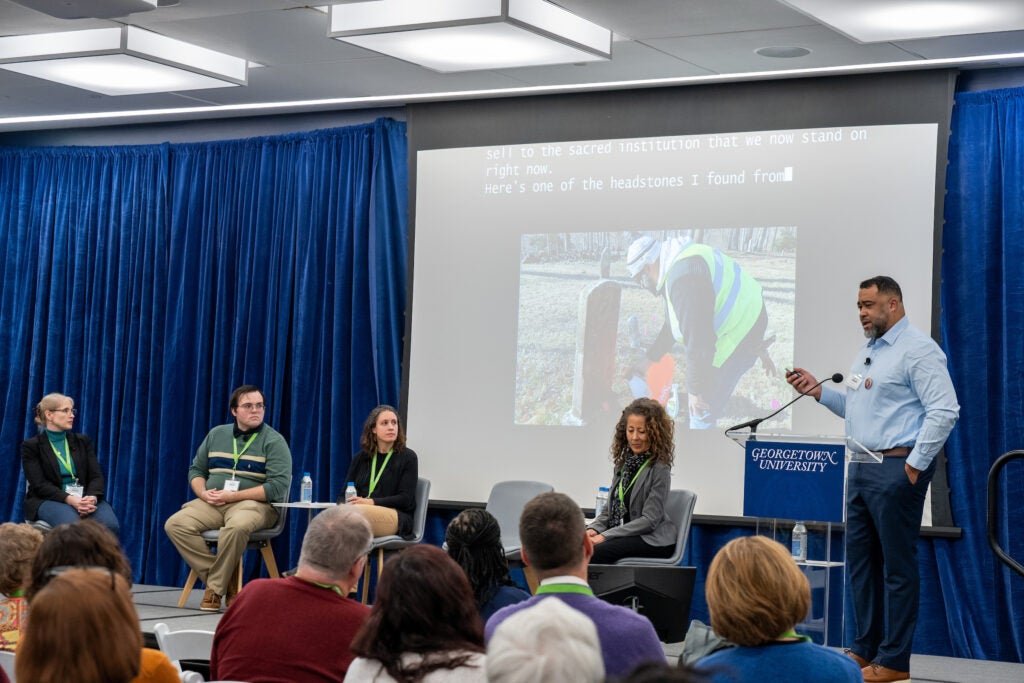 A black man speaks to a seated audience from behind a podium. Next to him on stage are four other people, seated as part of a discussion panel.