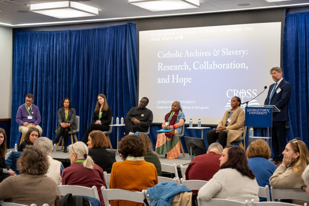 Adam Rothman, a white man, speaks to a seated audience from behind a podium. Next to him are six people of varying genders and races, seated as part of a panel discussion.