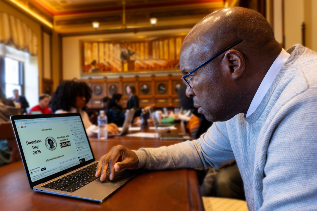 A black man sits at a wooden conference table in the Philodemic Room and leans over a laptop, whose screen is displaying an introduction slide for the Douglass Day 2026 broadcast.