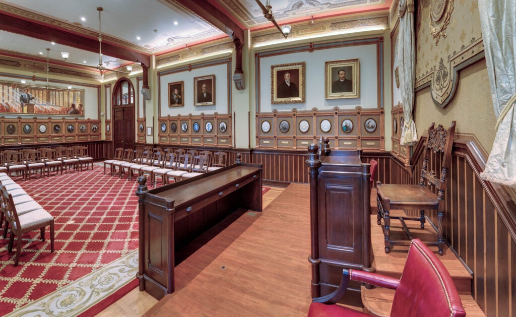 The Philodemic Room. The dark wood rostrum sits on a wooden stage in the foreground. The surrounding walls are covered in large and small portraits. Chairs line walls, surrounding a red and gray carpet.