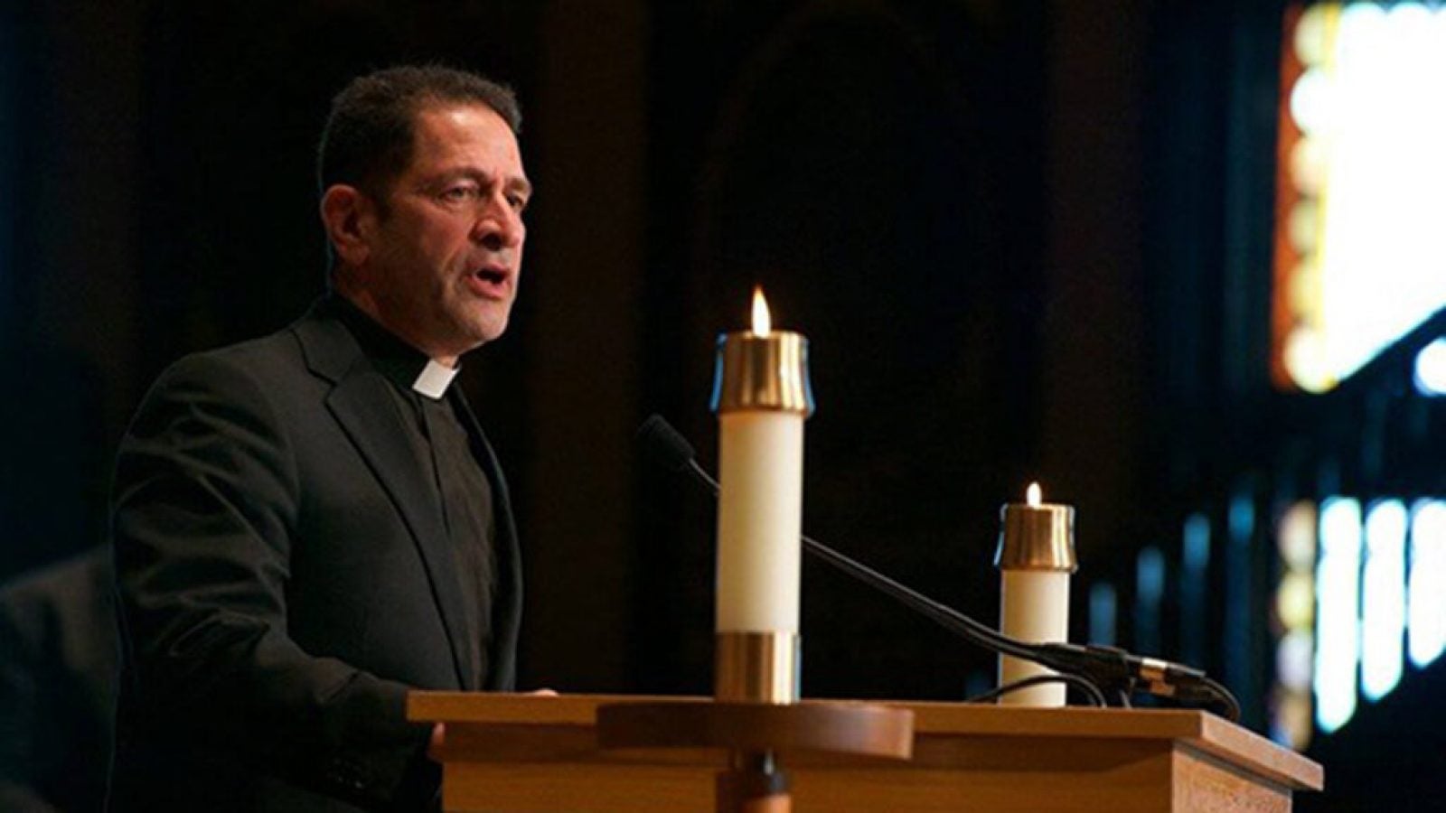 Fr. Kesicki, a white male minister speaks at from behind an mic'd altar with candles on either side.