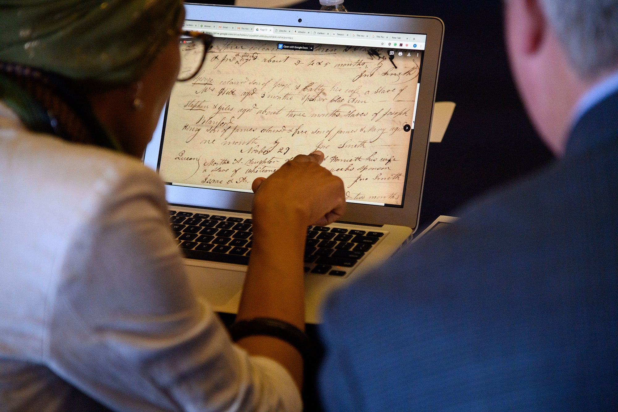 Two volunteers read a handwritten historical document on a laptop computer screen.